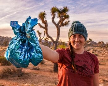 A woman volunteering to pick up trash in the desert holds two bags of collected trash out in front of her.