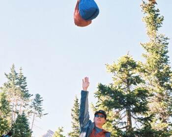 A man reaching up his right arm toward two food bags that are hung from a rope between two trees.